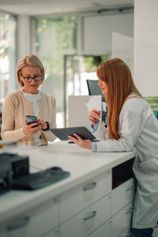 Pharmacist with tablet leaning on counter and selling drugs to old woman